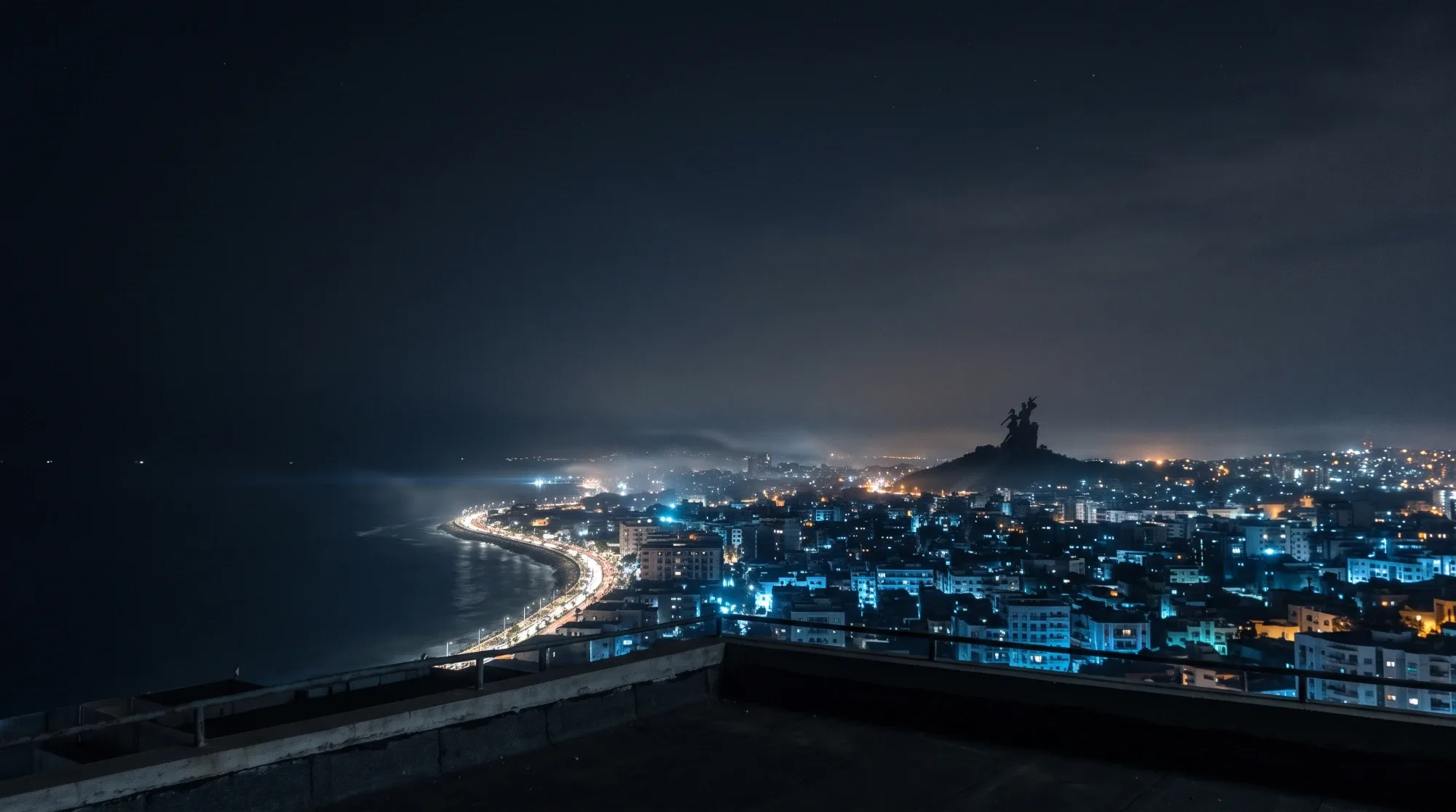 Dakar la nuit vue depuis un toit, la Corniche éclairée et le Monument de la Renaissance africaine sur sa colline